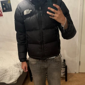 The North Face svart pufferjacka  - Säljer en svart pufferjacka från The North Face med ikonisk logga på bröstet och ryggen. Jackan har hög krage, dragkedja framtill och snörning i nederkant. En luva finns bakom dragkedja i kragen. Materialet är syntet och dun, perfekt för kalla dagar. Jackan är långärmad och har en lite cropped passform. Nypris: ca 3000kr