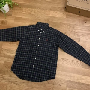 Rutig skjorta från Ralph Lauren XS - Snygg rutig skjorta från Ralph Lauren i mörkblått, grönt och rött. Skjortan har klassisk krage, långa ärmar och knappar framtill. Broderad röd logga på bröstet. Tillverkad i mjuk bomull som är skön mot huden. Perfekt för dig som gillar stilrena och tidlösa plagg.