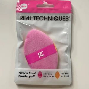 Real Techniques 2-i-1 powder puff - Miracle 2-i-1 powder puff från Real Techniques med en rosa sida för puder och en orange sida för flytande smink. Puffen har en squishy silikon-kärna och ett rosa band med RT-logga för extra kontroll. Perfekt för att blanda foundation, concealer och puder.