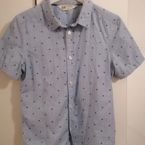 Blå kortärmad skjorta med stjärnor H&M - Snygg ljusblå skjorta från H&M med korta ärmar och små svarta stjärnor som mönster. Skjortan har klassisk krage, knappar framtill och är gjord i mjuk bomull. Perfekt för dig som gillar coola detaljer och enkel stil.