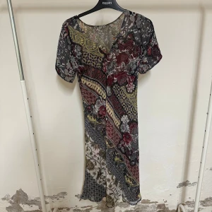 V-ringing klänning med fin print - Säljer en långklänning med v-ringning och korta ärmar. Klänningen har ett färgstarkt blommigt mönster i vinrött, grått, beige och inslag av gult. Materialet är polyester med ett coolt transparent uttryck. Perfekt för dig som gillar bohemisk stil och unika prints.