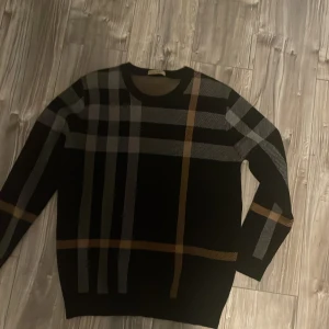 Rutig svart tröja från Burberry - Snygg svart tröja från Burberry med klassiskt rutigt mönster i grått och brunt. Rund halsringning och långa ärmar. Perfekt för dig som gillar stilrena designer med en lyxig touch. Materialet känns mjukt och skönt mot huden.