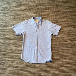 Blå kortärmad skjorta Tailored Originals - Fräsch ljusblå skjorta från Tailored Originals med klassisk krage och korta ärmar. Skjortan har knappar framtill och är i en normal passform. Perfekt för dig som gillar stilrena och enkla plagg. Materialet känns lätt och bekvämt mot huden.