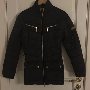 Svart quiltad pufferjacka Barbour - Svart pufferjacka från Barbour med quiltad design, guldfärgade dragkedjor och knappar, högt krage med spänne och bälte i midjan. Jackan har två bröstfickor med dragkedja och två fickor med knapp framtill. Perfekt för kalla dagar.