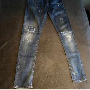 Dondup blå jeans med slitningar - Snygga Replay Anbass jeans i blå tvätt med slitna detaljer och hål på benen. Modellen har fem fickor, orange sömmar på bakfickorna och klassisk knappgylf. Jeansen är smala i benen och har en cool, lite ruffad look.