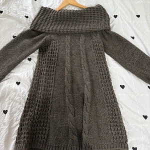 Brun stickad långklänning - Mysig brun stickad långklänning med bred krage och kabelstickade detaljer framtill. Klänningen har långa ärmar och ett chunky, strukturerat mönster som ger en cool och avslappnad vibe. Perfekt för kyliga dagar och att styla med boots.