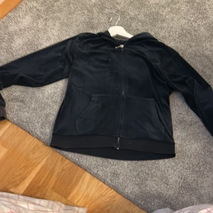 Svart velour hoodie från Juicy Couture - Mysig mörk blå hoodie från Juicy Couture i mjuk velour med dragkedja framtill och huva. Klassisk känguruficka och diskret broderad JC-logga på bröstet. Perfekt för chill dagar eller när du vill ha en skön och snygg tröja, frakt inkluderas i priset  