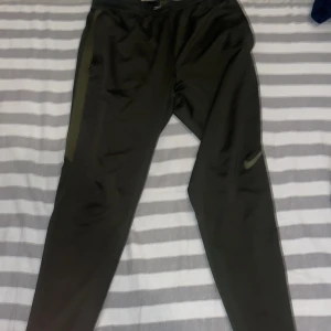 Gröna Nike träningsbyxor med grön logga - Säljer ett par svarta träningsbyxor från Nike med grön swoosh-logga på benet. Byxorna har smal passform, elastisk midja med snörning och gröna detaljer längs sidorna. Perfekta för träning eller chill, tillverkade i mjukt och stretchigt syntetmaterial.
