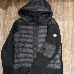 Svart Moncler pufferjacka - Svart pufferjacka från Moncler med stickade ärmar och huva. Jackan har quiltad front, dragkedja, fickor med knapp och en extra ficka med dragkedja på ärmen. Moncler-logga på vänster ärm. Perfekt för dig som gillar street och exklusiv stil.
