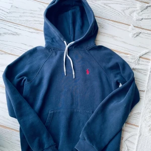 Marinblå hoodie från Polo Ralph Lauren - Säljer en marinblå hoodie från Polo Ralph Lauren med klassisk röd logga broderad på bröstet. Tröjan har huva med vita snören, känguruficka och långa ärmar. Perfekt för chill dagar och snygg streetstyle. Lite liten i storlek 