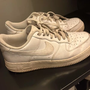 Nike Air Force 1 vita sneakers - Klassiska Nike Air Force 1 sneakers i helvitt skinn med rund tå och platt sula. Skorna har snörning och perforerade detaljer på tån för extra ventilation. Perfekt för dig som gillar streetstyle och vill ha en ikonisk sneaker i garderoben.