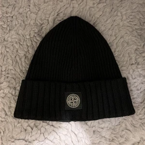 Svart ribbad mössa Stone Island Junior - Svart ribbstickad mössa från Stone Island Junior med uppvikt kant och klassisk logopatch framtill. Snygg och enkel design som funkar till streetwear och håller dig varm. Materialet är mjukt och stickat för extra komfort.