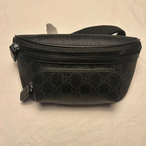 Svart Gucci magväska med GG-mönster - Snygg svart  Gucci magväska med klassiskt GG-mönster och justerbart band. Väskan har två dragkedjor med detaljer i skinn och en praktisk ficka framtill. Perfekt för att bära dina essentials och ge din outfit en lyxig touch.