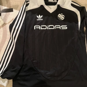 Svart långärmad Adidas fotbollströja XS - Säljer en svart långärmad fotbollströja från Adidas med vita detaljer och klassiska tre ränder längs ärmarna. Tröjan har en vit krage och logga på bröstet, samt ett unikt emblem. Materialet är lätt och andas, perfekt för träning eller match.