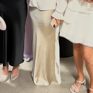 Champagnefärgad långkjol i satin - Säljer en elegant långkjol i champagnefärgad satin med lätt glansig finish. Kjolen har en figurnära passform upptill och faller snyggt mot benen. Endast använd en gång!