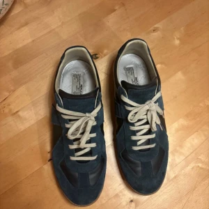 Maison Margiela sneakers blå - Maison Margiela sneakers i mörkblå mocka och skinn med beige snörning och gummisula. Klassisk low-top modell med rund tå och diskreta detaljer. Märkets logga syns på sulan och innersulan. Perfekta för dig som gillar stilrena och lyxiga sneakers.