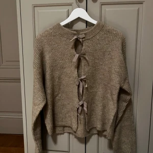 Beige stickad tröja med rosetter Gina Tricot - Superfin beige stickad tröja från Gina Tricot med unika rosettdetaljer längs ryggen. Tröjan har lång ärm, rund halsringning och en loose passform. Perfekt för höst och vinter när du vill vara både varm och snygg.