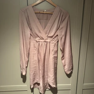 Ljusrosa omlottklänning med v-ringning MAMA - Snygg ljusrosa kortklänning med omlott-design och djup v-ringning. Klänningen har långa ärmar och ett lätt rynkat midjeparti som ger en feminin siluett. Materialet känns mjukt och har en lätt glans. Perfekt för dig som gillar stilrena och romantiska plagg. Passar bra under och efter graviditet och har använts som klänning och tunika.