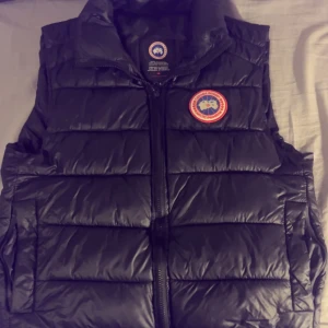 Svart dunväst från Canada Goose - Säljer en svart dunväst från Canada Goose med klassisk logga på bröstet. Västen har quiltad design, hög krage och dragkedja framtill. Perfekt för lager-på-lager och håller dig varm under kyliga dagar. Stilsäker och funktionell för dig som gillar streetwear.