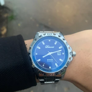 Blå och silverfärgad Doventi klocka - Snygg klocka från Doventi med blå urtavla och silverfärgat armband i metall. Klockan har datumvisning, markerade index och en sportig bezel med siffror. Quartz-urverk och klassisk rund form, perfekt för dig som gillar stilrena accessoarer.