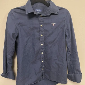 Mörkblå skjorta från GANT - Klassisk mörkblå skjorta från GANT med vit knappstängning och broderad logga på bröstet. Skjortan har lång ärm, krage och är tillverkad i mjuk bomull. Perfekt för dig som gillar stilrena och tidlösa plagg med snygg passform.