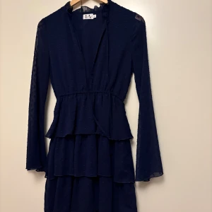 Marinblå volangklänning med knyt - Supersöt marinblå kortklänning från NA-KD med volanger i flera lager, transparenta långärmar och diskret prickigt mönster. Klänningen har v-ringning med knyt vid halsen och markerad midja för en snygg siluett.