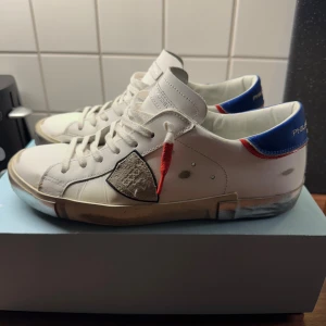 Philippe Model Prsx Low Leather Blue White  - Snygga vita Philippe Model sneakers i skinn med blå och röda detaljer på hälen, grå mockadetaljer och klassisk snörning. Skorna har en unik patch på sidan och coolt sliten look på sulan. Perfekta för dig som gillar streetstyle och fransk design. Pris går att diskutera vid snabb affär. Hör av er vid frågor 