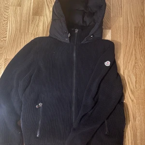 Svart Moncler pufferjacka med huva - Svart pufferjacka från Moncler med stickade ärmar och quiltad kropp. Jackan har en stor huva, dragkedja framtill och Moncler-logga på ärmen. Materialmix med stickad och syntet, perfekt för kyliga dagar. Snygg och modern look med ribbade detaljer och praktiska fickor.