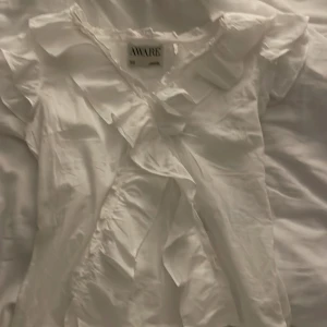 Vit blus med volang från AWARE - Säljer en vit blus från AWARE i mjukt bomullsmaterial. Blusen har korta ärmar och snygga volangdetaljer längs halsringningen och framtill. Perfekt för dig som gillar en feminin och fräsch stil. Passar dig som vill ha en lätt och luftig topp.