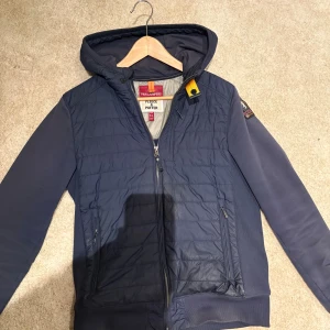 Blå cardigan från Parajumpers - Snygg mörkblå pufferjacka/cardigan från Parajumpers med huva och dragkedja. Cardiganen har quiltad framsida, två sidofickor med dragkedja och cool gul detalj vid halsen. Insidan är fodrad med grått tyg och har en säkerhetsficka. Perfekt för kalla dagar. Den är i storlek Y-L som är i herrstorlek S-M. Den är lite solblekt men inga andra defekter ✅ pris kan diskuteras 👍🏻💫