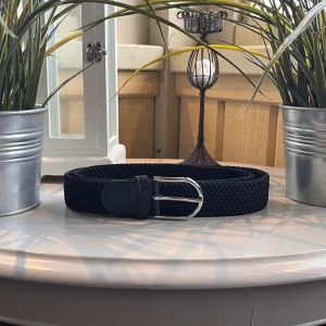 Marinblått flätat bälte med silverfärgat spänne - Snyggt marinblått flätat bälte med ett stilrent silverfärgat metallspänne. Bältet har en bred och mjuk design med detaljer i skinn vid spännet. Perfekt för att ge din outfit en clean och modern touch.