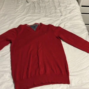 Röd v-ringad tröja Tommy Hilfiger - Röd v-ringad tröja från Tommy Hilfiger med broderad logga på bröstet. Tröjan har långa ärmar, ribbade muddar och är tillverkad i mjuk bomull. Perfekt för dig som gillar klassisk och stilren design. Inte använd