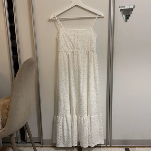 Vit långklänning från Chiquelle - Supersöt vit långklänning från Chiquelle med smala axelband och broderade spetsdetaljer. Klänningen har ett luftigt fall, volang nedtill och är tillverkad i bomull med ett blommigt hålmönster. Perfekt för sommarens alla tillfällen!