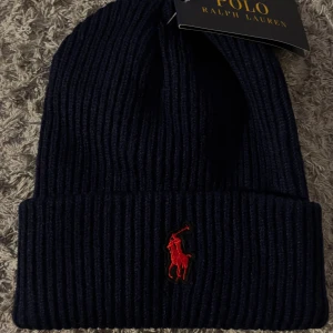 Mörkblå ribbstickad mössa Polo Ralph Lauren - Snygg mörkblå ribbstickad mössa från Polo Ralph Lauren med klassisk röd broderad logga framtill. Perfekt för kalla dagar och enkel att matcha med olika outfits. Tjockt uppvik för extra värme och stil.