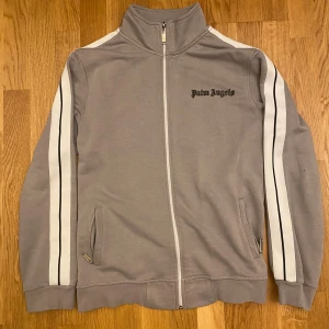 Grå track jacket från Palm Angels - Säljer en grå track jacket från Palm Angels med vita och svarta ränder längs ärmarna. Tröjan har hög krage, hel dragkedja framtill och två fickor med dragkedja. Klassisk streetwear-look med logga på bröstet.