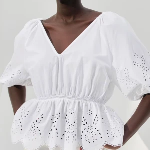 Vit blus med broderade detaljer - Säljer en vit blus med v-ringning och puffiga korta ärmar. Blusen har broderade hålmönster och volang nedtill, vilket ger en romantisk och fräsch vibe. Perfekt för dig som gillar ljusa och luftiga toppar med fina detaljer.