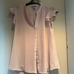 Ljusrosa blus med volangärm från KappAhl - Säljer en ljusrosa blus från KappAhl med korta volangärmar och v-ringad hals. Framsidan har snygga vertikala veck och knappar hela vägen. Blusen är tillverkad i ett tunt, flowy material som ger en luftig känsla. 