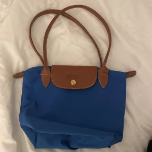 Blå axelväska - Snygg blå axelväska från Longchamp med bruna skinnhandtag och lock. Väskan har guldfärgad knapp och dragkedja upptill. 