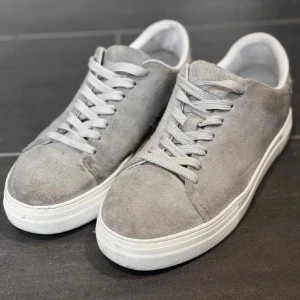 Grå mockasneakers från Selected Homme - Snygga grå sneakers från Selected Homme i mocka med vit platt sula och klassisk snörning. Skorna har en stilren och minimalistisk design som passar perfekt till jeans eller chinos. Tillverkade i Portugal och riktigt sköna på foten.