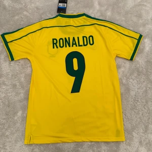 Brasilien Ronaldo 9 Nike fotbollströja - Gul Brasilien fotbollströja från Nike med gröna detaljer, nummer 9 och Ronaldo på ryggen. Klassisk design med gröna ränder längs axlarna, broderat landslagsmärke och Nike-logga på bröstet. Tillverkad i lätt och ventilerande material.