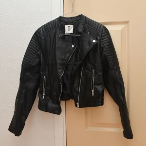 Svart skinnjacka från H&M, strl 164 - Snygg svart skinnjacka från H&M med coola quiltade detaljer på axlar och ärmar. Jackan har asymmetrisk dragkedja framtill, två zip-fickor och en kortare, slim passform. Perfekt för dig som gillar en edgy stil och vill ha en jacka som sticker ut.