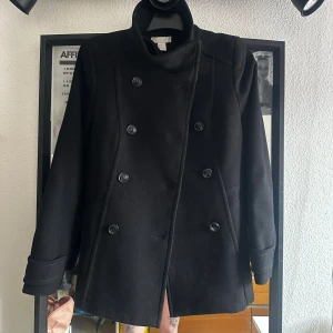 Svart kappa med dubbelknäppning - Säljer en svart kappa från H&M med snygg dubbelknäppning och hög krage. Jackan har långa ärmar, klassisk rak passform och är tillverkad i ullblandning. Perfekt för dig som gillar stilrena och tidlösa plagg med lite edge.