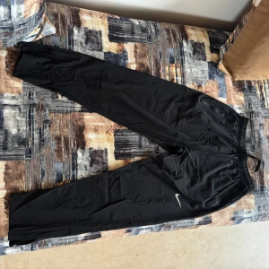 Svarta Nike träningsbyxor med snörning - Svarta träningsbyxor från Nike med elastisk midja och snörning. Byxorna har två fickor med dragkedja och en vit Nike-logga på benet. Materialet är lätt och syntetiskt, perfekt för sport eller chill. Passformen är normal och benen är raka.