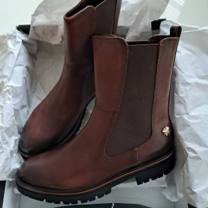 Bruna chelsea boots fr. Marco Tozzi  - Snygga bruna chelsea boots i skinn med grov platt sula och rund tå. Baksidan har ett coolt krokodilmönster och bootsen är dekorerade med en liten guldfärgad blomdetalj. Elastiska paneler på sidorna gör dem enkla att ta på. Perfekta för höst och vinter.