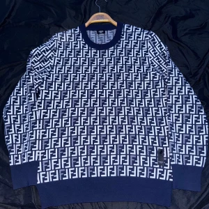 Blåvit stickad Fendi tröja - Säljer en blåvit stickad tröja från Fendi med ikoniskt FF-mönster över hela plagget. Tröjan har rund halsringning, långa ärmar och ribbade muddar. Perfekt för dig som vill sticka ut med en lyxig och trendig look.