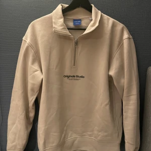Beige half zip tröja Jack & Jones XS - Snygg beige tröja från Jack & Jones Originals med half zip och hög krage. Mjuk insida och tryck med 'Originals Studio' på bröstet. Perfekt för dig som gillar enkel och clean stil. Tillverkad i bomullsmix för skön känsla.