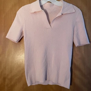 Ljusrosa ribbad polotopp - Säljer en ljusrosa ribbad polotopp med korta ärmar och snygg krage. Toppen har en v-ringad öppning framtill och är gjord i ett mjukt, stickat material som känns skönt mot huden. Perfekt för dig som gillar stilrena och enkla plagg med en twist.