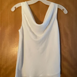 Assymetrisk Vit ärmlös linnetopp med draperad hals - Säljer en stilren vit linnetopp med snygg draperad halsringning. Toppen är ärmlös och har en mjuk, flowy passform som ger en chill och fräsch vibe. Perfekt för varma dagar eller när du vill ha en clean look. Materialet känns lätt och svalt mot huden.