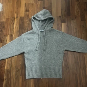 Grå stickad hoodie från Zara - Mysig grå hoodie från Zara i stickat material. Tröjan har huva med snören, ribbade muddar och bred ribbad nederkant. Perfekt för kyliga dagar och enkel att matcha med jeans eller mjukisbyxor.