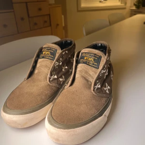 Bruna WTAPS x Vans sneakers med broderi - Unika bruna sneakers från WTAPS x Vans med broderade vita korslagda ben på sidorna. Skorna har platt sula, rund tå och detaljer i mocka och canvas. Klassisk Vans-gummisula och metallöglor för snörning. Perfekt för dig som gillar streetwear och coola samarbeten. Pris ej hugget i sten!